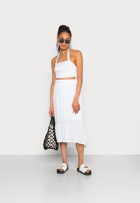 Top curto branco com alças, combinado com uma saia midi branca com bainha em babados. Os acessórios incluem óculos de sol pretos e uma mala preta com padrão.