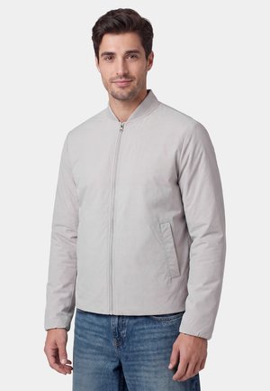 Mand iført en lysegrå lynlåstrøje og blå jeans, står mod en ensfarvet lys baggrund og kigger på kameraet.