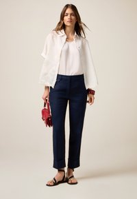 Blusa bianca leggera con dettagli decorativi, abbinata a pantaloni navy su misura, sandali neri e una piccola borsa rossa.