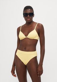 Yellow ribbed bikini set featuring a triangular bralette with thin straps and high-waisted bottoms. Model wears oversized black sunglasses.