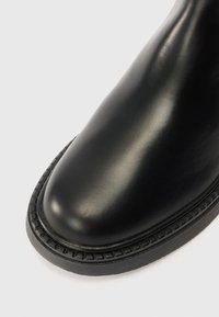 Crne kožne čizme s okruglim vrhom, glatkom površinom i teksturiranom gumenom potplatom s uzdignutim, patterned rubom oko ivice.