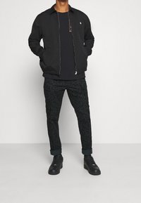 Svart bomberjacka med elastiska muddar, svart t-shirt och svarta mönstrade byxor. Klädseln kompletteras med svarta sneakers.