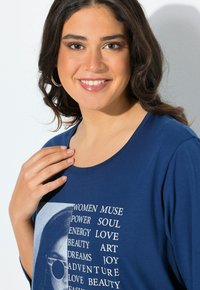 Granatowa koszulka z okrągłym dekoltem. Posiada graficzny wzór twarzy oraz biały tekst z wyrazami takimi jak "KOBIETY", "MOC" i "PRZYGODA."