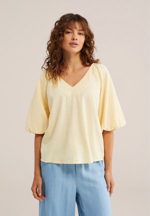 Kobieta o kręconych włosach w jasnżółtej bluzce z dekoltem w serek i bufiastymi rękawami oraz jasnoniebieskich spodniach, stojąca na jednolitym tle.