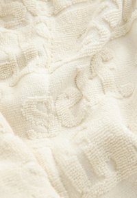 Tissu crème texturé avec une surface douce et pelucheuse. Présente des motifs en relief, offrant de la profondeur et un intérêt visuel. Apparence légèrement froissée.