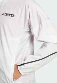 Giacca bianca con accenti in rete, striscia nera sulla manica e branding Terrex. Texture liscia con dettagli a motivo geometrico.