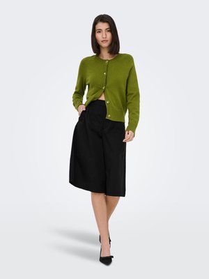 Frau steht und trägt eine olivgrüne, geknöpfte Strickjacke, schwarze, weite, knielange Shorts und schwarze spitze Absätze vor weißem Hintergrund.