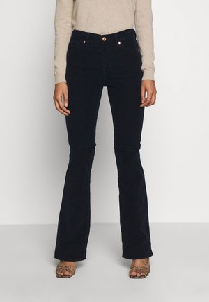 Person trägt hoch taillierte schwarze Schlaghose, beiges Langarmshirt und offene Sandalen mit Absatz und Knöchelriemen vor einfachem Hintergrund.