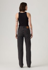 Woman with short brown hair wearing a black sleeveless top and high-waisted dark gray straight-leg jeans, standing facing away on a plain background.