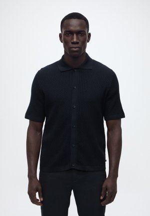 Mann mit dunkler Hautfarbe, der ein schwarzes kurzärmeliges Strickhemd mit Knöpfen und schwarze Hosen trägt, steht vor einem schlichten hellen Hintergrund.