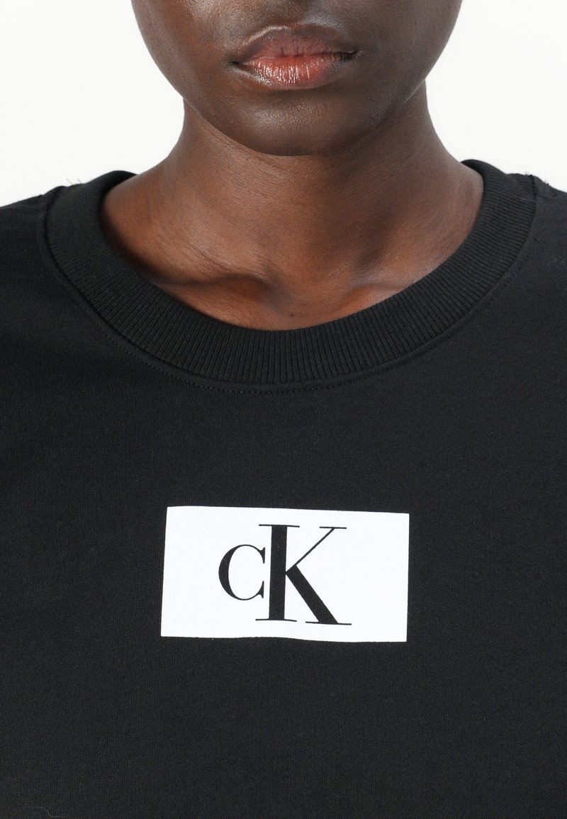 Sudadera negra con un logotipo rectangular blanco que presenta "cK" en letras negras en negrita. Diseño de cuello redondo con textura acanalada en el cuello.
