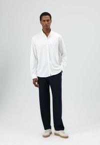 Weißes, langärmliges Hemd mit Knöpfen, kombiniert mit marineblauer Hose und weißen Turnschuhen. Glattes Material mit entspanntem Schnitt und klassischem Kragen.