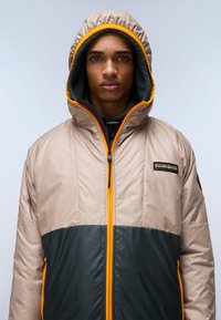 Tan en donkergroene geïsoleerde jas met oranje accenten, voorzien van een volledige rits, een capuchon en een logo-patch op de borst. Gladde textuur.