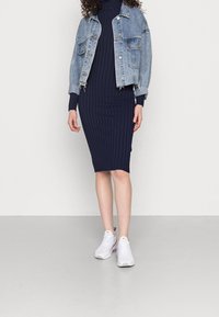 Femme portant une robe côtelée bleu marine, une veste en jean courte et des baskets blanches, debout sur un sol gris devant un mur blanc.