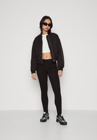 Chaqueta bomber negra, top corto blanco, jeans negros de cintura alta y zapatillas negras con detalles amarillos. Modelo luciendo gafas de sol.