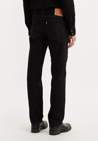Personne portant un jean noir droit avec un patch en cuir de marque et une languette rouge, associé à une chemise noire et des chaussures en cuir noires, debout.