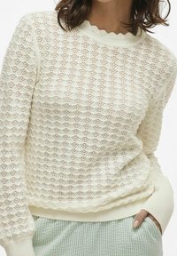 Pull léger en crème avec un motif en maille en losanges, un col à bord scallopé et des poignets côtelés. Associé à un pantalon à carreaux vert et blanc.