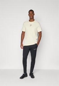 Světle béžové tričko s malým červeným a černým logem, kombinované s černými slim-fit tepláky s bílým logem. Model má na sobě černé boty.