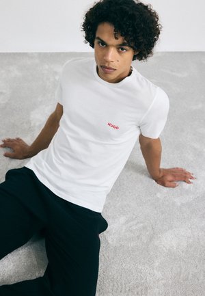 Weißes Baumwoll-T-Shirt mit rundem Ausschnitt, versehen mit einem roten "HUGO"-Logo auf der linken Brust. Das Modell trägt schwarze Hosen und sitzt auf einer hellen, strukturierten Oberfläche.