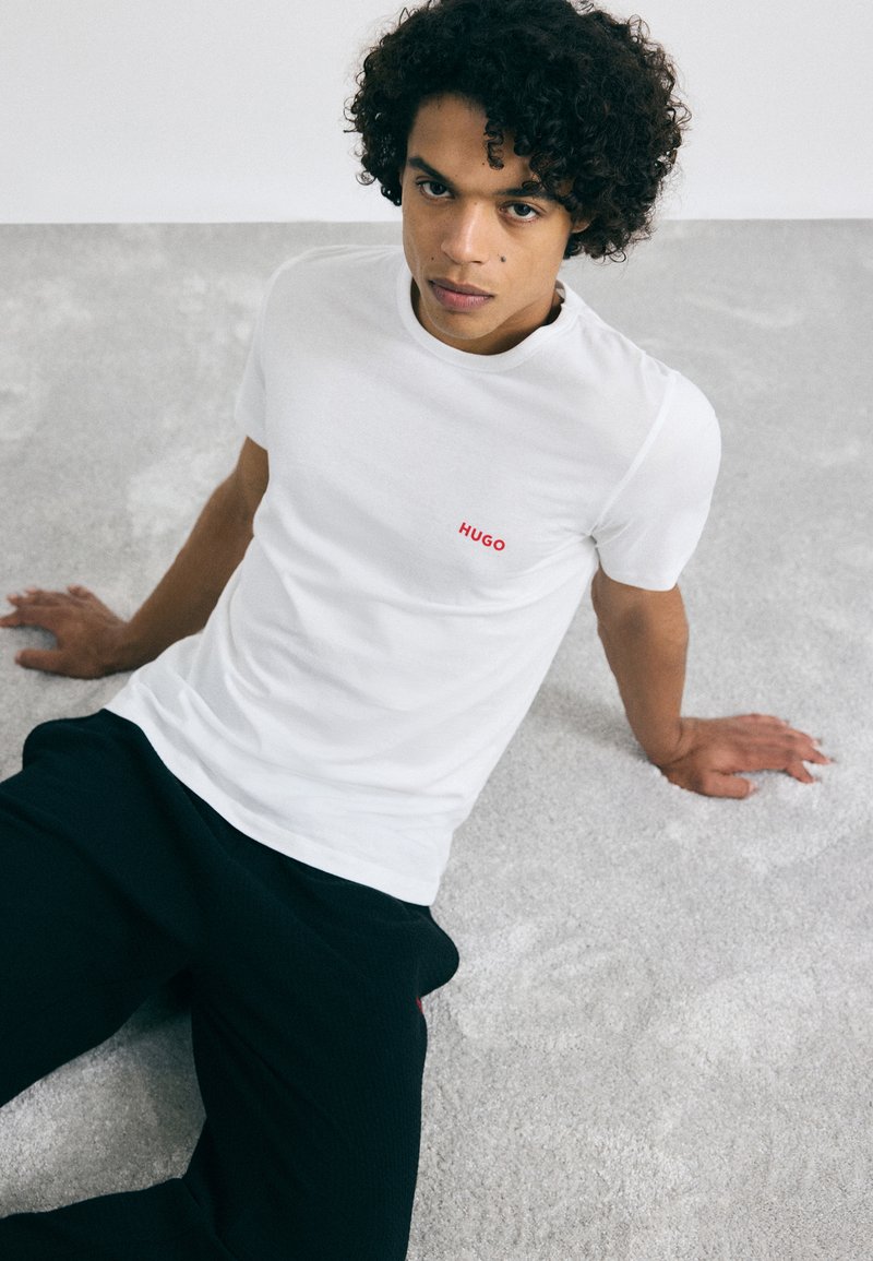 T-shirt en coton blanc avec un col rond, présentant un logo rouge "HUGO" sur la poitrine gauche. Le modèle porte un pantalon noir, assis sur une surface texturée claire.