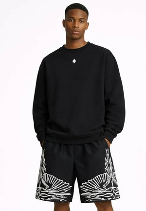 Ung mand iført en sort sweatshirt med et lille hvidt logo og sorte shorts med et stort hvidt abstrakt vingedesign.