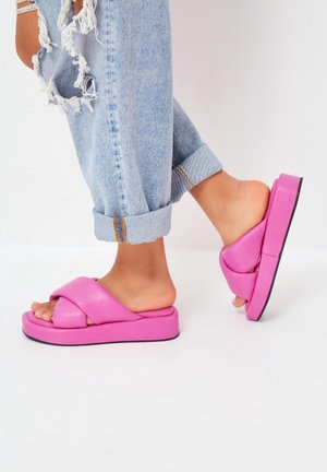 Lyserøde faux læder slidesandaler med en tykk platform, der har et kryds-rem design og en kontrasterende sort sål, parret med rullede denimbukser.