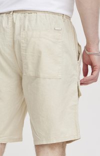 Un short cargo beige possède une taille élastique, des poches latérales et une poche arrière. Le tissu semble léger avec une texture douce.