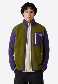 Giacca in pile con blocchi di colore verde e viola, tasche laterali e sul petto con cerniere, colletto e una texture morbida.