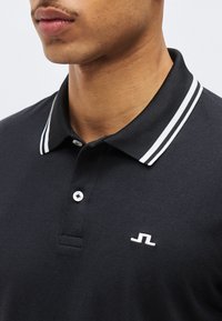 Κοντινό πλάνο ενός ατόμου που φοράει μαύρο πόλο με γιακά διπλής λευκής ρίγας και μικρό λευκό κεντημένο λογότυπο στο στήθος.