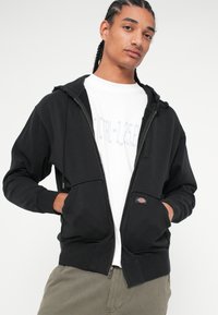 Μαύρο φούτερ με φερμουάρ και μπροστινή τσέπη, με ριμπ μανσέτες. Φορεμένο πάνω από μία λευκή μπλούζα, που αναδεικνύει ένα λογότυπο και έχει χαλαρή εφαρμογή.