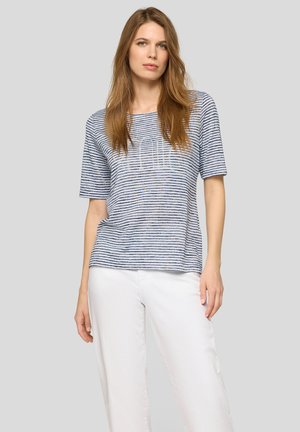 Woman with long light brown hair wearing a short-sleeve blue and white striped top and white pants standing against a gray background.