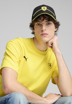 Yellow cotton t-shirt with Puma logo, black cap with yellow accents and Ferrari logo, model in casual pose, blue jeans.