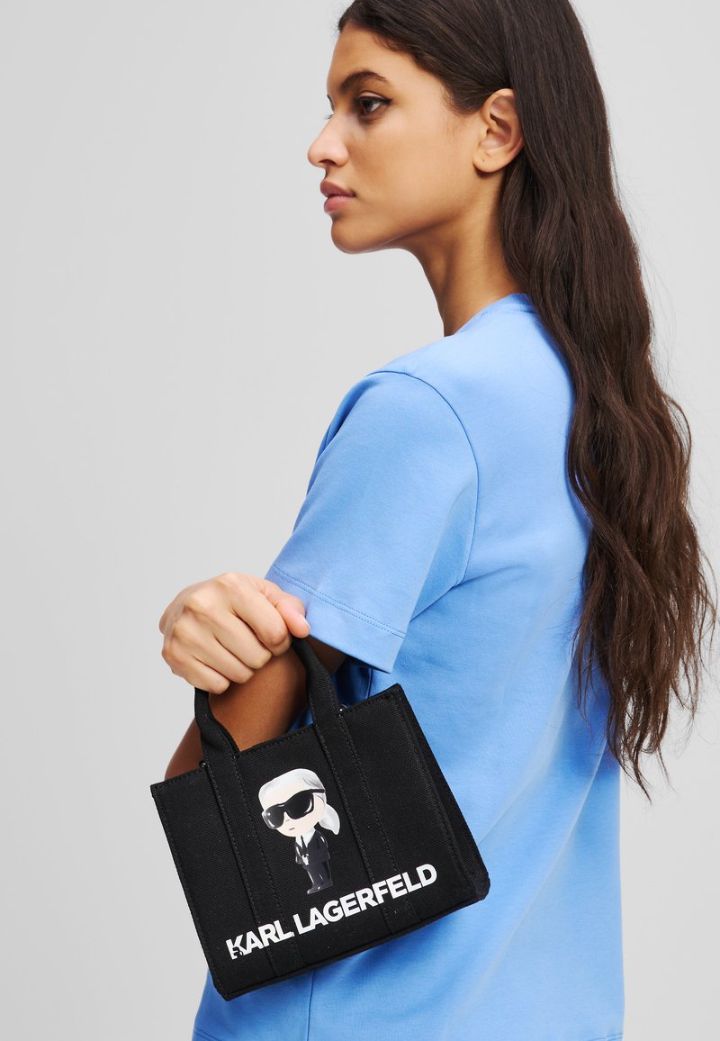Sac fourre-tout noir en toile arborant un design de dessin animé d'une figure portant des lunettes de soleil et un costume, avec "KARL LAGERFELD" imprimé en blanc.