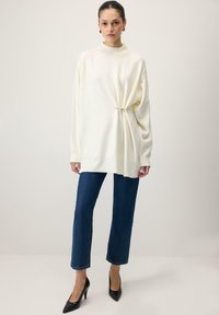 Kreemjas oversize-suurusega kampsun kõrge kaelusega, külgedelt tõmmatud detailidega, kombineeritud tumeliste sirge säärega teksapükste ja mustade terava ninaga kõrgete kontsadega.