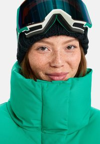 Turquoise puffer jacket with high collar, featuring a smooth texture. Black knitted beanie and ski goggles with mirrored lens and white accents.