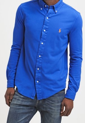 Mężczyzna w jasnoniebieskiej koszuli zapinanej na guziki z małym pomarańczowym logo na piersi, w ciemnoniebieskich dżinsach, stojący na jednolitym tle.