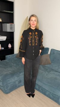 Žena stojí v interiéri oblečená v čiernej košeli s oranžovou kvetinovou výšivkou, čiernych džínsoch a čiernych špicatých topánkach pri modrej kordovej pohovke.