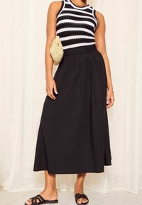 Černá maxi sukně s elastickým pasem, spárovaná s černobílým proužkovaným topem bez rukávů. Doplňky zahrnují tkanou béžovou kabelku.