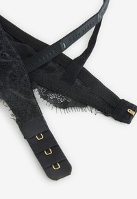 Čierna čipková podprsenka s krížovými ramienkami, so sieťovinovým materiálom, zlatými kovovými detailmi a textúrovaným povrchom. Nastaviteľné zapínanie na háčiky a očká.