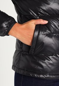 Sort pufferjakke med en blank overflade, elastiske manchetter og en sidelomme, med et quiltet design og glat tekstur.