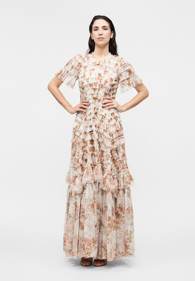 Γυναίκα που στέκεται με τα χέρια στους γοφούς, φορώντας μακρύ, στρωματοποιημένο φλοράλ φόρεμα με κοντά βολάν μανίκια σε απλό λευκό φόντο.