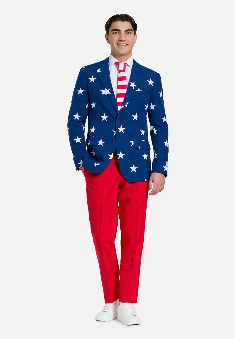 Blå blazer med hvide stjerner, røde bukser og et stribet slips. Hvide sneakers fuldender outfittet, som har et levende og dristigt design.