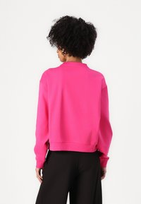 Felpa fucsia con design cropped, polsini e colletto a coste. Tessuto morbido, presenta una vestibilità rilassata. Pantaloni neri visibili sotto.