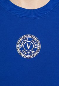 Blauwe katoenen trui met een ronde, goudkleurige geborduurde logo met de tekst "VERSACE JEANS COUTURE" en een kettinkjesontwerp. Geribbelde halslijn detail.