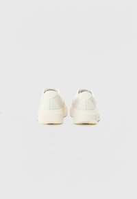 White sneakers with textured fabric upper, rounded shape, cushioned sole, and subtle logo detail on the back. Smooth finish and light color scheme.