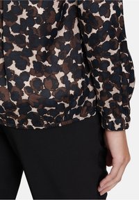 Leopardenmuster Bluse mit elastischen Bündchen und Saum, getragen mit schwarzen Hosen, zeigt eine Person mit der rechten Hand entspannt an ihrer Seite.