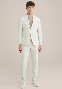 Weißer Anzug mit strukturiertem Finish, Einknopfverschluss, Reverskragen, passenden Hosen und hellgrauem Unterhemd, kombiniert mit weißen Sneakers.