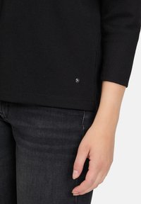 Mano femminile appoggiata accanto a una camicia nera a maniche lunghe texturizzata e a dei jeans grigio scuro, su uno sfondo bianco.