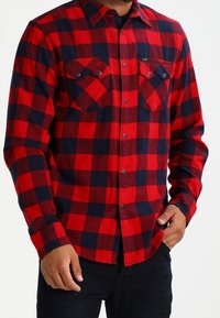 Camisa de franela a cuadros en rojo y azul marino con mangas largas, frente de botones y dos bolsillos en el pecho con botones de metal y una etiqueta de la marca.