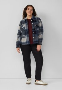 Karierte Wolljacke in Marineblau, Creme und Grau; mit Knopfverschluss und zwei Brusttaschen. Kombiniert mit einem kastanienbraunen Pullover und schwarzen Hosen. Weiße und braune Sneakers.
